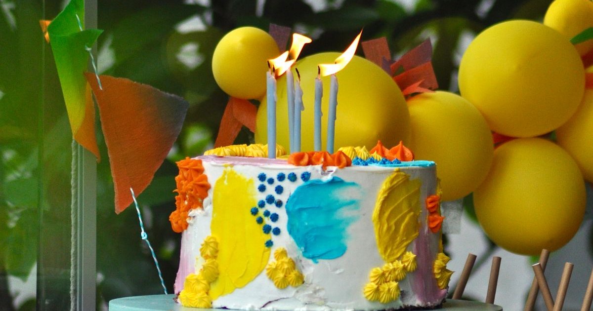 Bolo de aniversário decorado com velas acesas e balões amarelos ao fundo